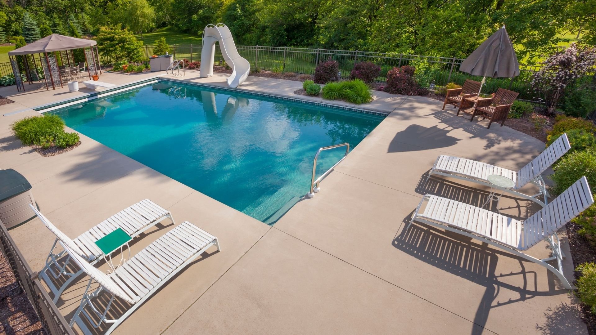 Luxury backyard pool with lounge chairs, waterslide, and surrounding green landscaping
